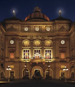 Den oplyste facade p&aring; Det Ny Teater i K&oslash;benhavn med en stor kuppel, s&oslash;jler og runde vinduer. Indgangen er centralt placeret og flankeret af dekorative detaljer.
